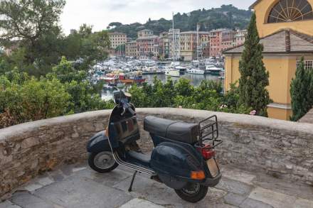 Classic Vespa parked overlooking a picturesque harbor in Italy, with colorful buildings and boats.